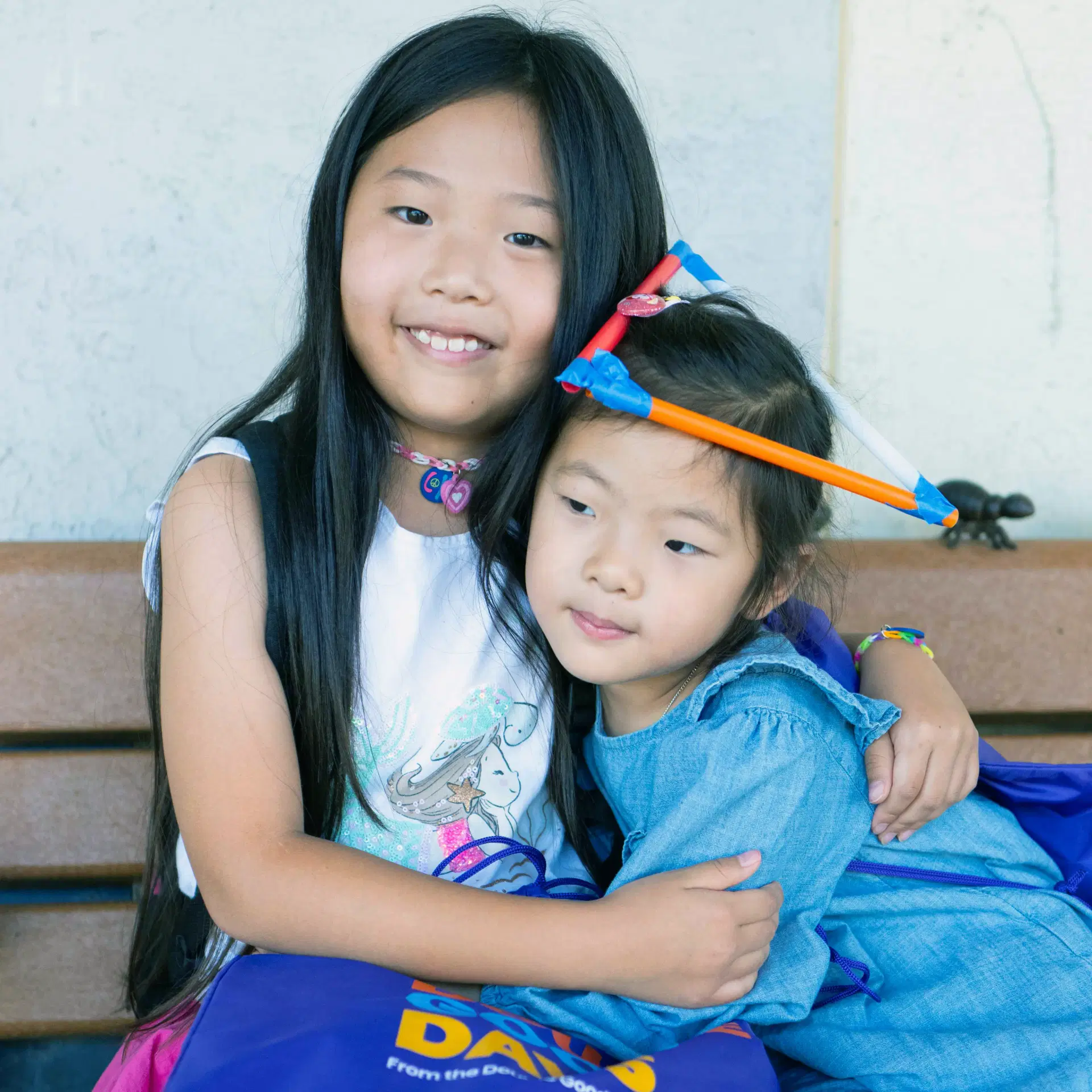 Two girls hugging each other while sitting on a bench. One is looking at the camera and the other is looking away.