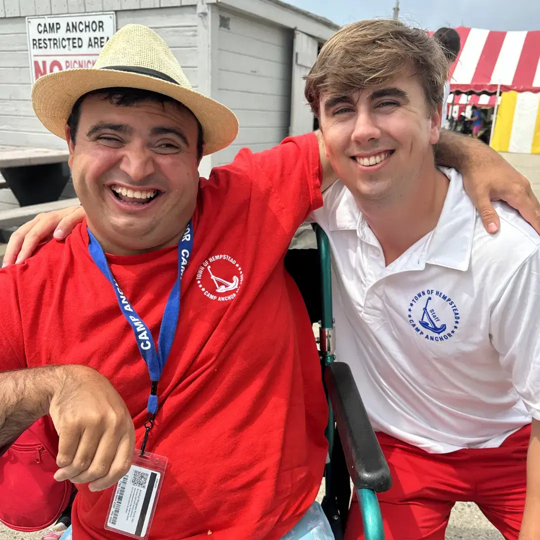 Two smiling men at Camp Anchor. One wears a red shirt and straw hat, the other a white shirt. A wheelchair visible between them.