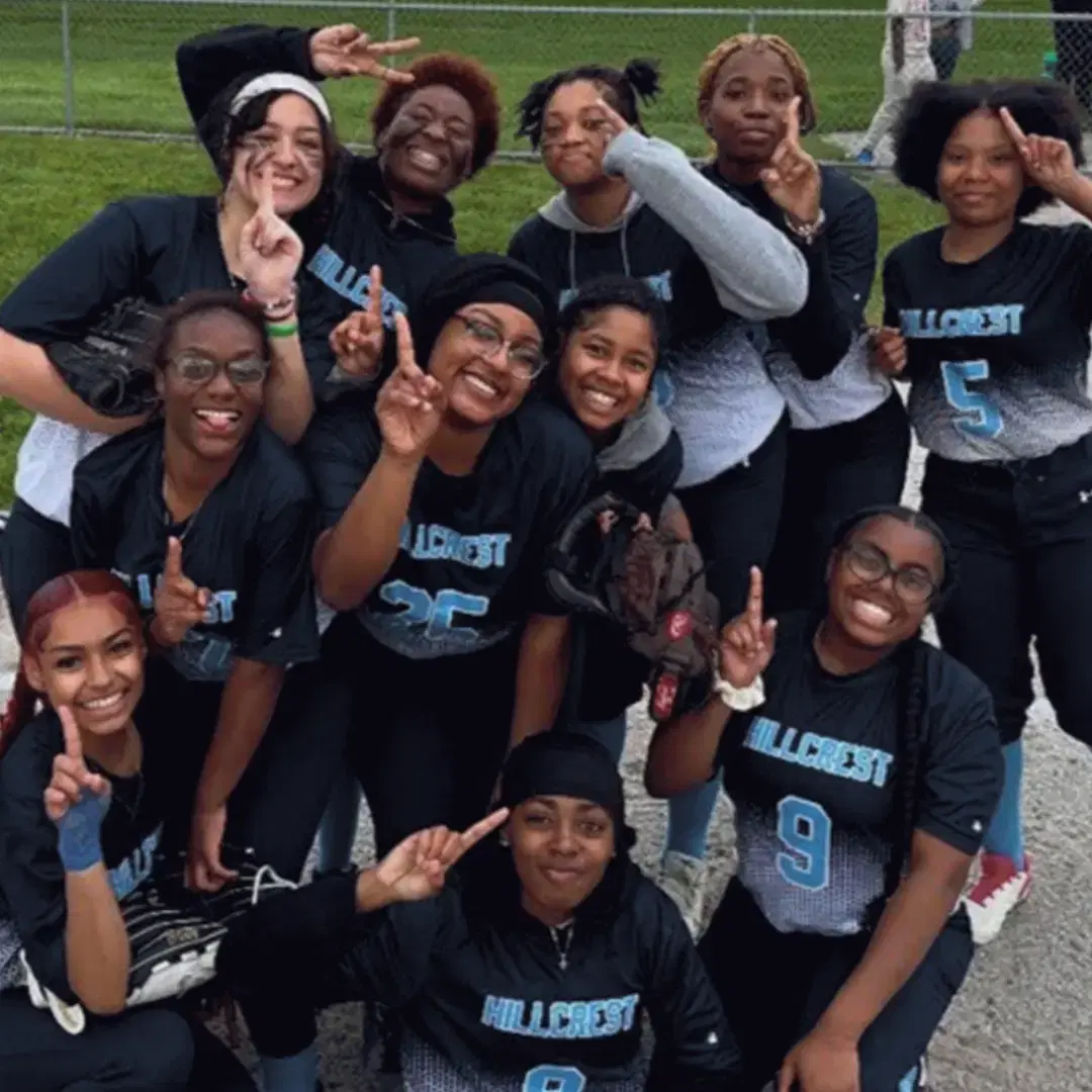 A joyful softball team in black "Hillcrest" jerseys poses on a field, smiling and making peace signs.